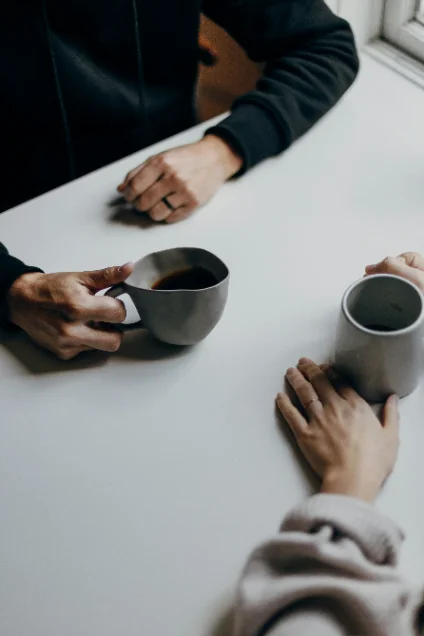 Two people sitting across a table from each other, holding mugs. Their faces are not shown.
